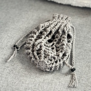 May include: A gray crocheted drawstring bag with black beads on the cords. The bag is lying on a gray textured surface.