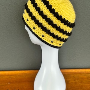 May include: A handmade yellow and black crochet beanie hat. The hat features horizontal black stripes and small black beads. The hat is displayed on a white mannequin head. The logo "fox creations" is visible in the bottom left corner.