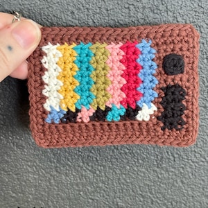 May include: Handmade crocheted keychain in the shape of a retro television. The screen shows a colourful test pattern in white, yellow, teal, pink, red, and blue. The TV frame is brown, with a black knob and antenna.