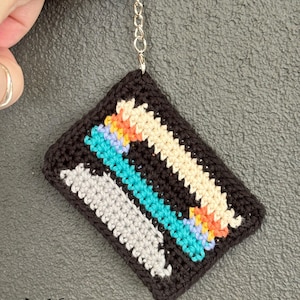 May include: A handmade, crocheted keychain in the shape of a cassette tape. The black keychain features colorful stripes in cream, turquoise, gray, orange, and purple. The keychain has a silver chain and ring. The text "fox creations" is visible.
