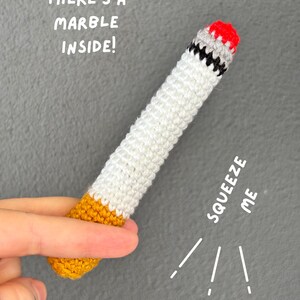 May include: A cylindrical crochet toy resembling a cigarette with a yellow filter, a white body, a black band, and a red tip. The text "THERE'S A MARBLE INSIDE!" and "SQUEEZE ME" are printed on the image. The item features a textured yarn construction.