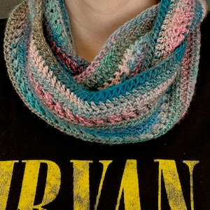 Peut inclure: Une écharpe infinie au crochet présentant un motif de fil panaché avec des nuances mélangées de bleu sarcelle, rose, marron et crème. L'écharpe a un motif de points côtelés texturés et est portée autour du cou sur un t-shirt noir avec du texte jaune.