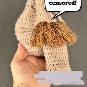Puede incluir: Una bolsa de ganchillo beige con cordón que presenta un bigote de lana marrón sujeto en la parte delantera. Un bocadillo blanco sobre el bigote contiene el texto negro "I've been censored!". La bolsa tiene una textura de hilo anudado.