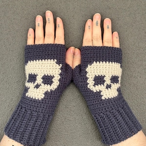 Puede incluir: Un par de guantes sin dedos de color gris carbón oscuro tejidos a ganchillo con un motivo de calavera beige claro en el dorso de cada mano. Los guantes presentan un diseño de puño acanalado y un orificio para el pulgar, con el texto fox creations.