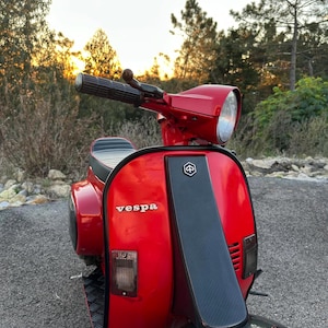 Op de afbeelding: Een klassieke rode Vespa scooter met zwarte accenten, geparkeerd op een verharde ondergrond. De scooter heeft het Vespa-logo en een opvallende koplamp. De achtergrond omvat bomen en een zonsondergang.