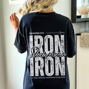 May include: Black t-shirt with the text "PROVERBS 27:17 IRON Sharpens IRON SO ONE FRIEND SHARPENS ANOTHER" in white. The shirt has a distressed, vintage look. The design includes a sword graphic above the text.