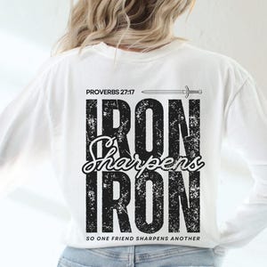 May include: White long-sleeve shirt with the text "PROVERBS 27:17" and "IRON SHARPENS IRON" in black. A sword graphic is above the text. The shirt is worn with light blue jeans.