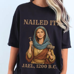 Könnte beinhalten: Schwarzes T-Shirt mit dem Text "NAILED IT." über einer Illustration einer Frau, die einen Hammer und einen Nagel hält. Der Text "JAEL, 1200 B.C." steht darunter. Die Frau trägt eine blaue Kopfbedeckung und ein goldfarbenes Gewand.