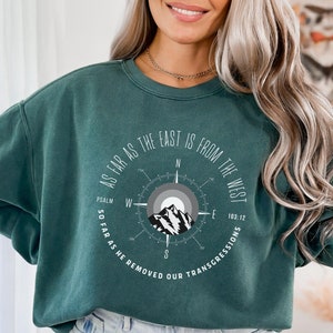May include: Dark teal sweatshirt featuring a compass design with a mountain graphic at the center. The text "As far as the East is from the West" and "So far as he removed our transgressions" are printed around the compass.