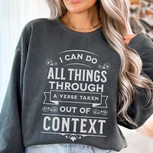 May include: Dark gray sweatshirt with the text "I CAN DO ALL THINGS THROUGH A VERSE TAKEN OUT OF CONTEXT" in white. The sweatshirt has a crew neck and long sleeves. The text is in a decorative font.