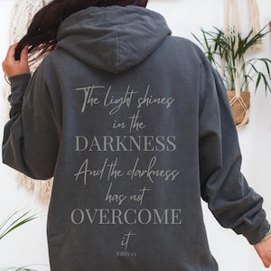 Puede incluir: Sudadera con capucha gris oscuro con el texto "The light shines in the DARKNESS And the darkness has not OVERCOME it JOHN 1:5" impreso en la espalda. La sudadera tiene una capucha con cordón y un bolsillo de canguro.
