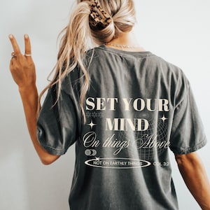 以下が含まれることがあります： クリーム色の「SET YOUR MIND On things Above」の文字が入った濃いグレーのTシャツ。 ゆったりとしたフィット感で、金髪の人が着ています。 テキストには「NOT ON EARTHLY THINGS COL 3:2」も含まれています。