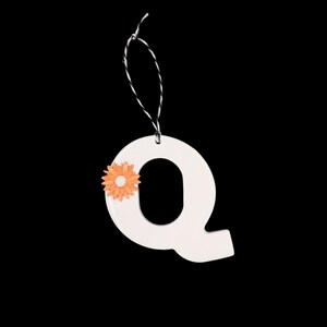 May include: White ceramic letter "Q" ornament with an orange flower accent. The ornament hangs from a black and white string against a black background. A decorative item for home decor or crafting.