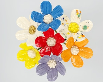 Könnte beinhalten: Eine Ansammlung bunter Keramikblumen in verschiedenen Farbtönen, darunter Blau, Rot, Gelb, Orange, Weiß und Lila. Jede Blume hat eine weiße Mitte. Die Blumen sind zusammen angeordnet und bilden eine lebendige und fröhliche Darstellung.