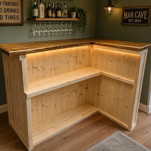 May include: An L-shaped wooden home bar featuring a dark stained countertop and light natural wood paneling. The interior includes two tiers of shelving and integrated LED strip lighting along the underside of the bar top edge.