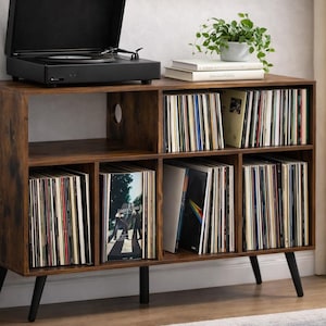 May include: A brown wooden record player stand with black legs. The stand has multiple compartments filled with vinyl records. A black record player sits on top, with a small potted plant and books.