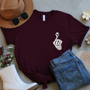May include: A maroon t-shirt with a white skeleton hand forming a heart shape, with a small heart above the fingers. The shirt is styled with blue jeans, a brown hat, sunglasses, and white boots. The shirt is made of a soft material.