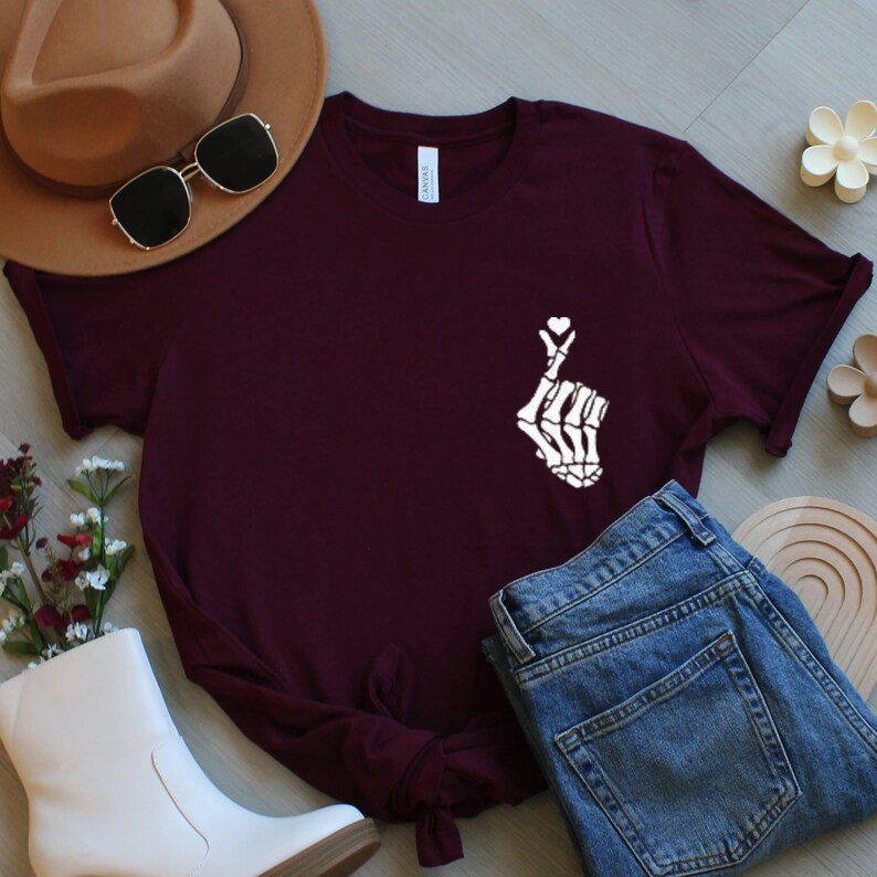May include: A maroon t-shirt with a white skeleton hand forming a heart shape, with a small heart above the fingers. The shirt is styled with blue jeans, a brown hat, sunglasses, and white boots. The shirt is made of a soft material.