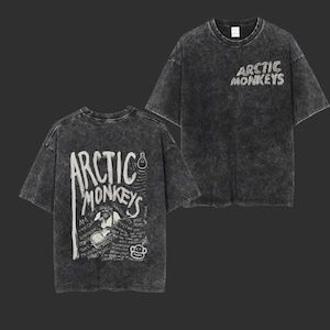 Pu&ograve; includere: Due viste di una t-shirt grigio antracite, lavata con acido. La parte anteriore presenta il nome della band "Arctic Monkeys" in un carattere bianco consumato. Il retro della maglietta presenta una grafica con il nome della band, un disegno di una persona che suona la chitarra e altro testo.