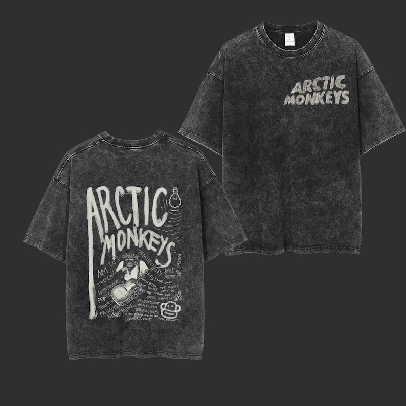 Pu&ograve; includere: Due viste di una t-shirt grigio antracite, lavata con acido. La parte anteriore presenta il nome della band "Arctic Monkeys" in un carattere bianco consumato. Il retro della maglietta presenta una grafica con il nome della band, un disegno di una persona che suona la chitarra e altro testo.