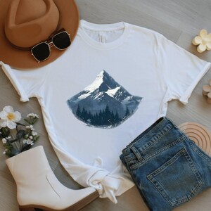 May include: White t-shirt featuring a mountain graphic with a snowy peak and a forest at the base. The shirt is paired with blue jeans, a brown hat, sunglasses, and white boots. A floral arrangement and wooden accents complete the casual ensemble.