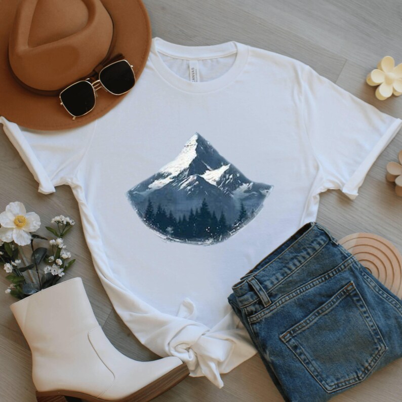 May include: White t-shirt featuring a mountain graphic with a snowy peak and a forest at the base. The shirt is paired with blue jeans, a brown hat, sunglasses, and white boots. A floral arrangement and wooden accents complete the casual ensemble.