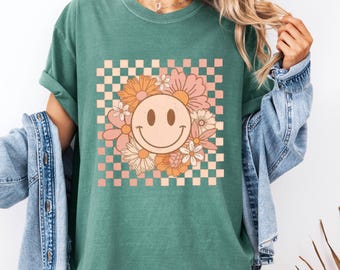 May include: Green t-shirt featuring a smiley face surrounded by flowers in shades of pink, peach, and white, set against a checkered background. The shirt has rolled-up sleeves.