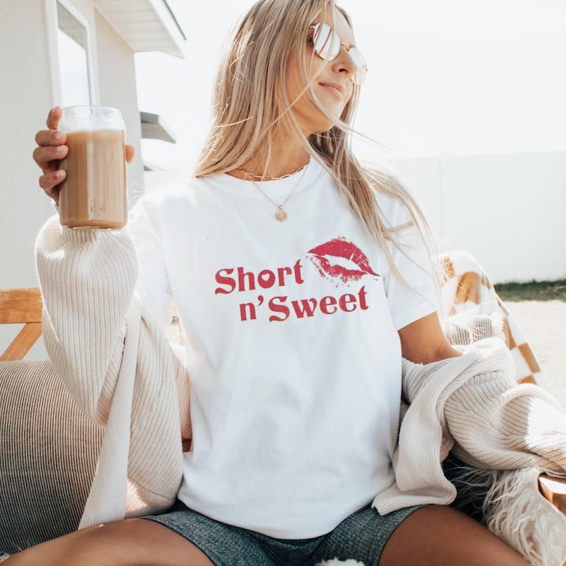 May include: White t-shirt with the text "Short n' Sweet" in red script, and a red lip print. The person is holding a glass of iced coffee. The person is wearing sunglasses and a gold necklace.