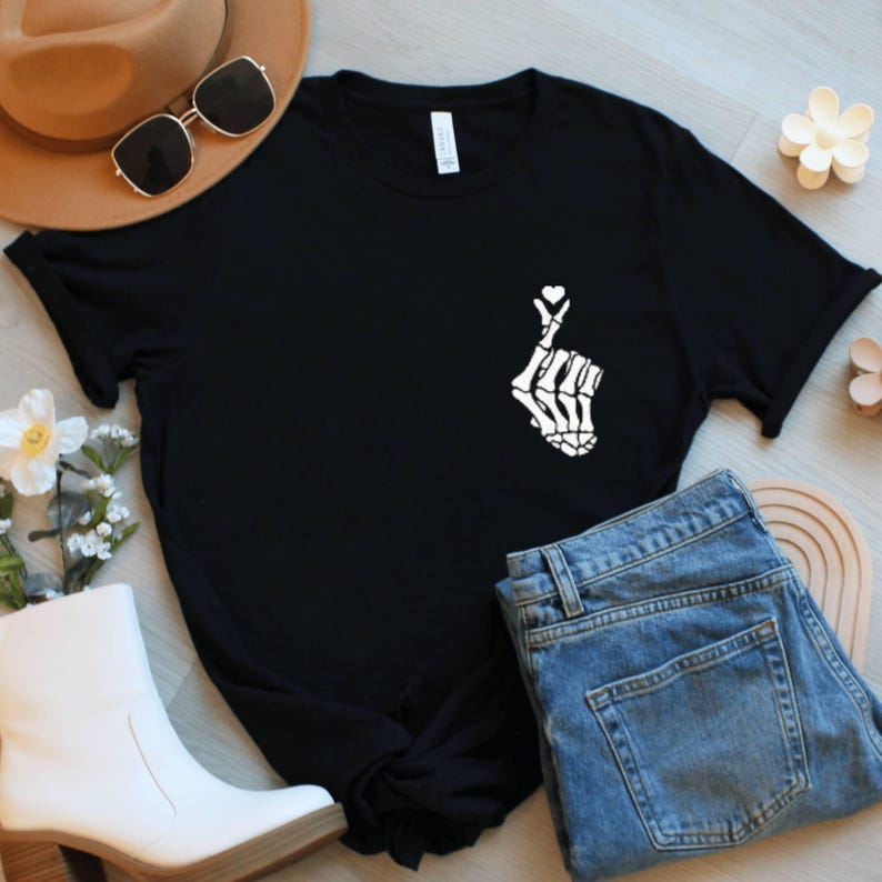 May include: Black t-shirt featuring a white skeleton hand forming a heart shape. The shirt is styled with blue jeans, a brown hat, sunglasses, and white boots. Decorative flowers and accessories are also visible.