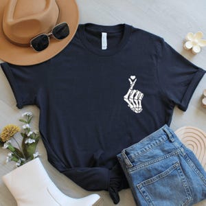 May include: A black t-shirt with a white skeleton hand forming a heart shape. The image includes blue jeans, a brown hat, and sunglasses. A white boot and floral decorations are also visible, creating a casual style.
