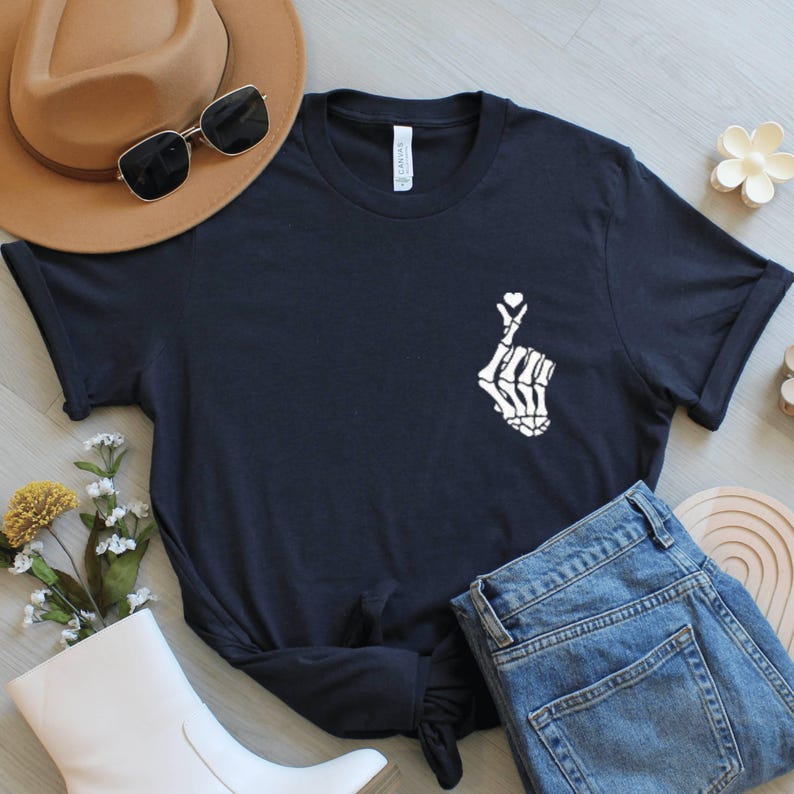 May include: A black t-shirt with a white skeleton hand forming a heart shape. The image includes blue jeans, a brown hat, and sunglasses. A white boot and floral decorations are also visible, creating a casual style.