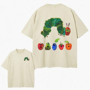 May include: A beige short-sleeve t-shirt featuring a small green caterpillar graphic on the front left chest. The back displays a large illustration of a green caterpillar and a row of five colorful fruits including an apple, pear, plum, strawberry, and orange.