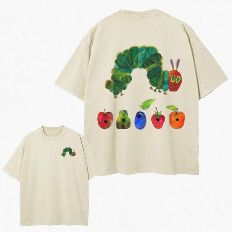 May include: A beige short-sleeve t-shirt featuring a small green caterpillar graphic on the front left chest. The back displays a large illustration of a green caterpillar and a row of five colorful fruits including an apple, pear, plum, strawberry, and orange.