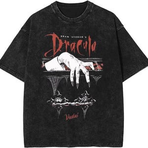 May include: Black t-shirt featuring a graphic from Bram Stoker's Dracula. The design includes a white hand with long black fingernails reaching out, with the word "Dracula" in red script. The shirt also has the word "Vudu!" printed on it.