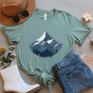 May include: Sage green t-shirt featuring a mountain graphic with a snow-capped peak and a forest base. The shirt is styled with a brown hat, sunglasses, white boots, and blue jeans. A casual, nature-themed outfit.