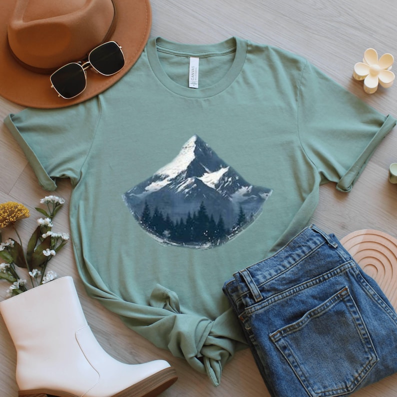 May include: Sage green t-shirt featuring a mountain graphic with a snow-capped peak and a forest base. The shirt is styled with a brown hat, sunglasses, white boots, and blue jeans. A casual, nature-themed outfit.