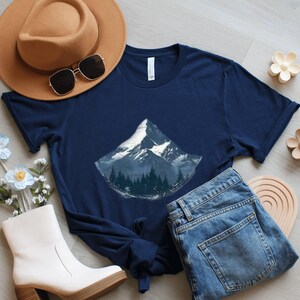 May include: Navy blue t-shirt featuring a mountain graphic with a snow-capped peak and a forest at the base. The shirt is styled with a brown hat, sunglasses, white boots, and blue jeans. The shirt is made of a soft material.