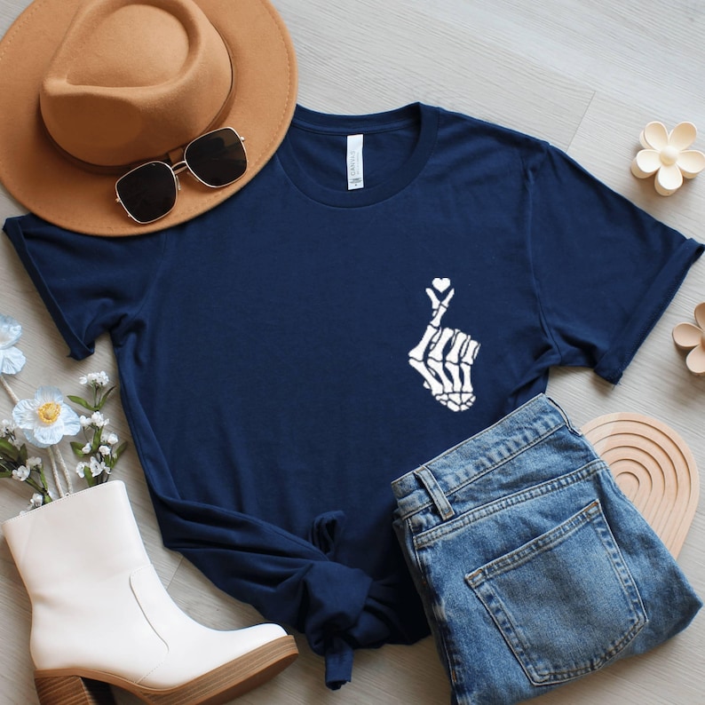 May include: A navy blue t-shirt featuring a white skeleton hand forming a heart shape. The shirt is styled with blue denim jeans, a brown hat, sunglasses, and white ankle boots. Decorative flowers and a wooden coaster are also visible.