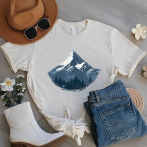 May include: Cream-colored t-shirt featuring a mountain graphic with a snowy peak and a forest base. The shirt is paired with blue jeans, a brown hat, sunglasses, and white boots, suggesting a casual, outdoorsy theme.
