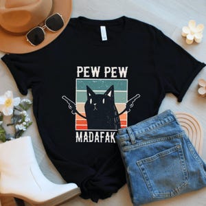 May include: Black t-shirt featuring a cartoon cat with guns and the text "PEW PEW MADAFAK". The design includes a retro color block background with horizontal stripes in teal, green, orange, and red. The shirt is paired with blue jeans.