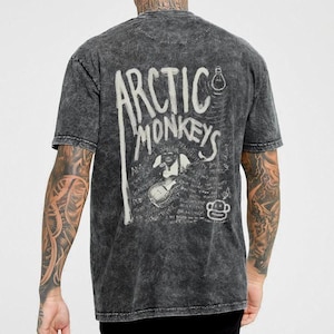 Pu&ograve; includere: T-shirt grigio scuro con il nome della band "Arctic Monkeys" in bianco, insieme a una grafica di una scimmia che suona la chitarra. La maglietta ha un aspetto vintage e usurato.