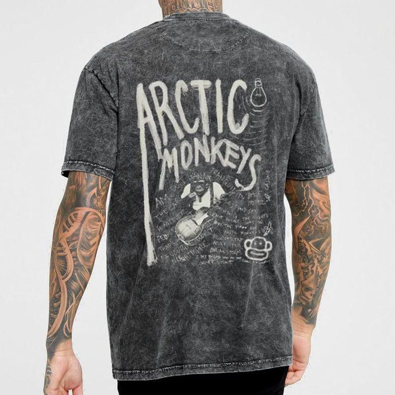 Pu&ograve; includere: T-shirt grigio scuro con il nome della band "Arctic Monkeys" in bianco, insieme a una grafica di una scimmia che suona la chitarra. La maglietta ha un aspetto vintage e usurato.