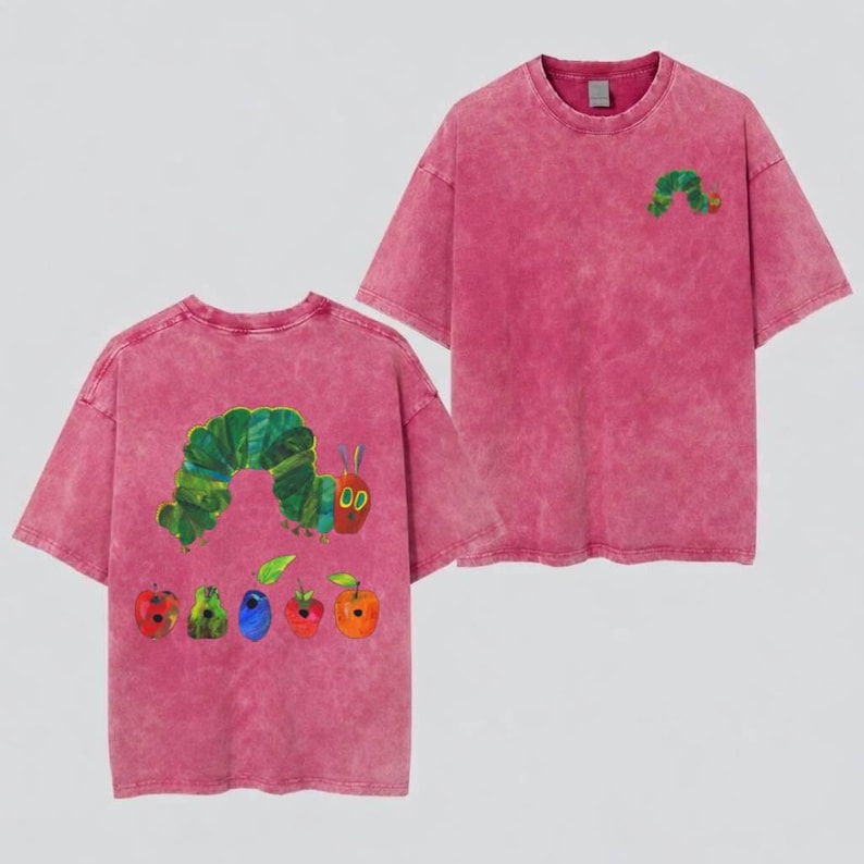 May include: A mineral-washed pink t-shirt featuring a small green caterpillar graphic on the front chest. The back displays a large illustration of a green caterpillar alongside various colorful fruits including apples, pears, and plums in a textured style.