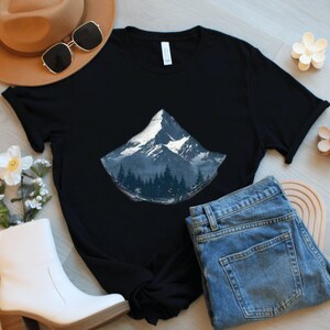 May include: Black t-shirt featuring a mountain graphic with a snow-capped peak and a forest at the base. The shirt is paired with blue jeans, a brown hat, sunglasses, and white boots. The design is a nature-themed graphic.