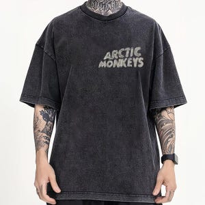 Pu&ograve; includere: T-shirt grigio scuro con la scritta "ARCTIC MONKEYS" in un font strutturato. La maglietta ha maniche corte e girocollo. Il tessuto ha un aspetto sbiadito e vintage.