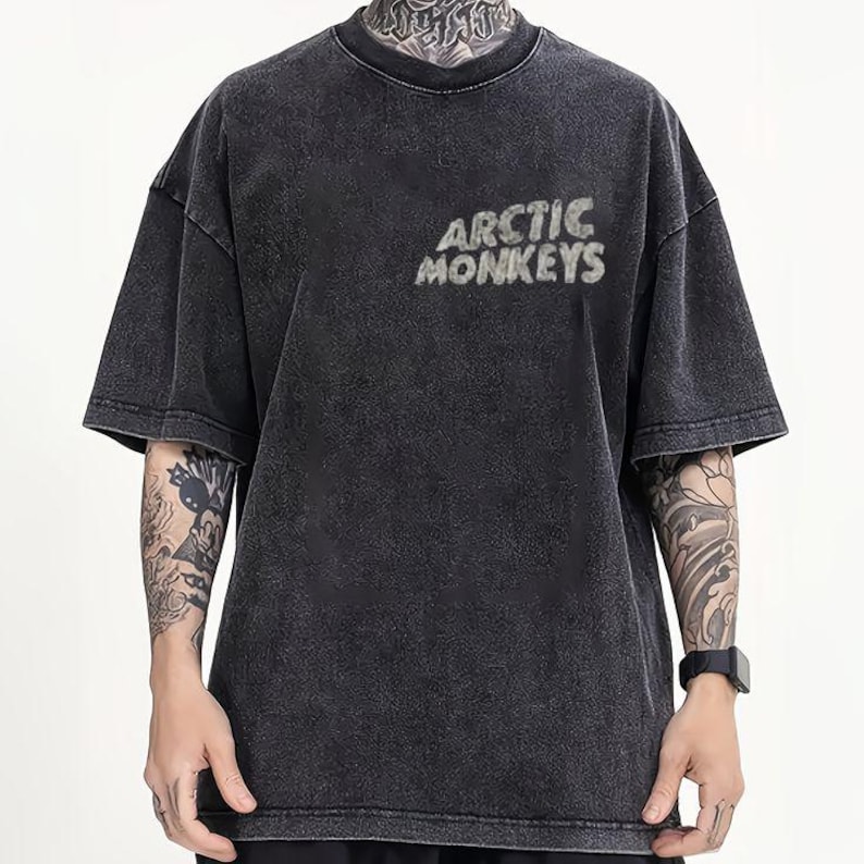 Pu&ograve; includere: T-shirt grigio scuro con la scritta "ARCTIC MONKEYS" in un font strutturato. La maglietta ha maniche corte e girocollo. Il tessuto ha un aspetto sbiadito e vintage.