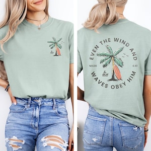 May include: A sage green t-shirt featuring a small palm tree and surfboard graphic on the front left chest. The back displays a large circular design with a palm tree, a surfboard, and the text "EVEN THE WIND AND WAVES OBEY HIM MARK 4:41".