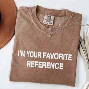 May include: A brown Comfort Colors t-shirt with the text "I'M YOUR FAVORITE REFERENCE" in white. A silver necklace with a small pendant rests on the shirt. A brown hat and dried palm leaf are in the background.