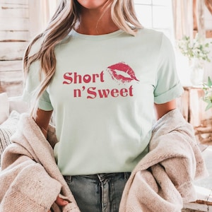 May include: A pale green t-shirt featuring the words "Short n' Sweet" in red, accompanied by a red lip print. The shirt has rolled-up sleeves and is made of a soft fabric. The model is wearing blue jeans and a beige cardigan.