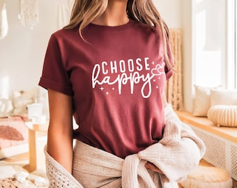 May include: A maroon t-shirt with the words "CHOOSE happy" in white script. The design includes stars and hearts. The shirt is being worn by a person.